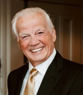 Older man with white hair wearing a black blazer, white dress shirt, and tan tie, smiling at the camera.
